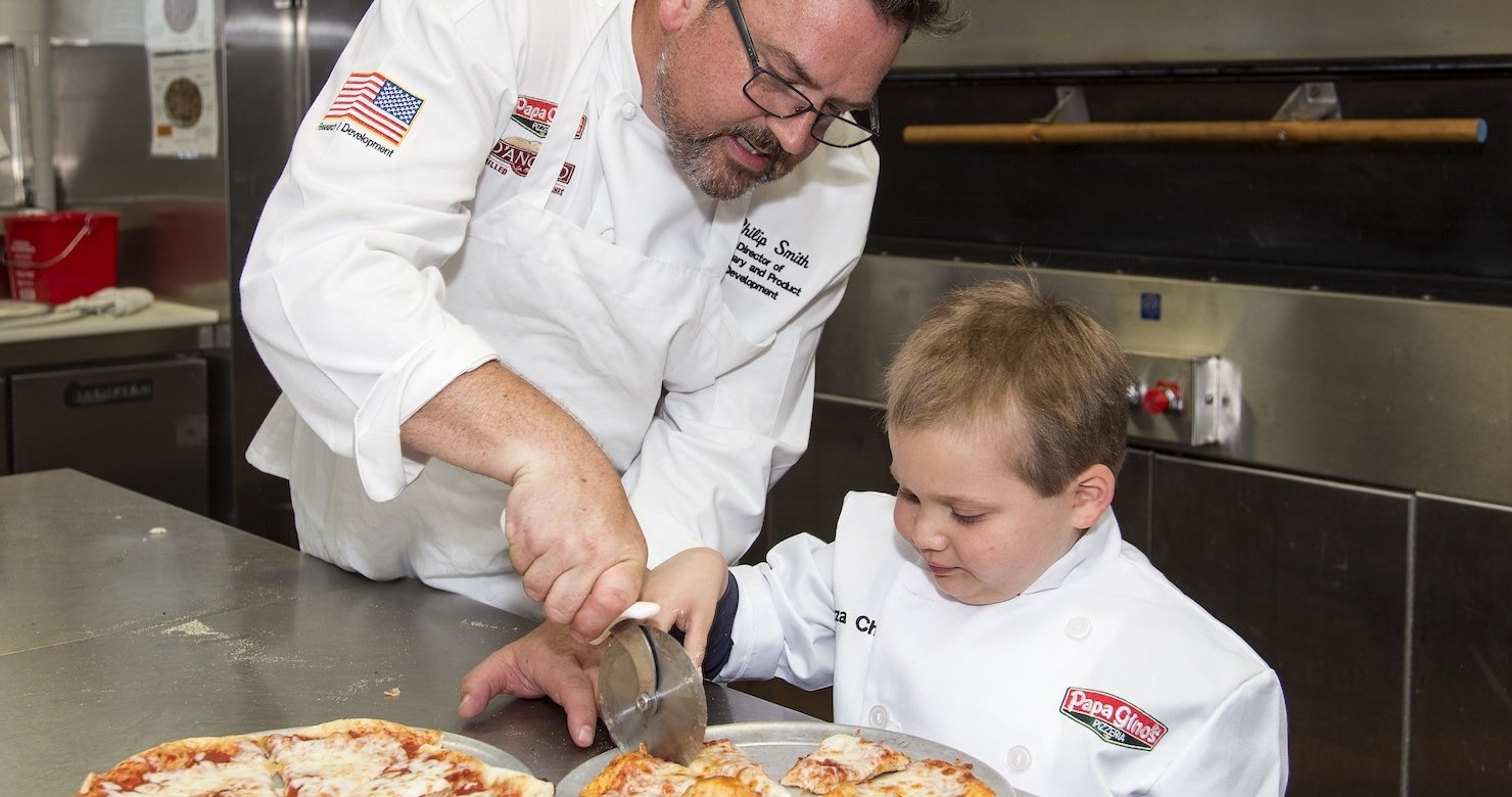 Young patient in chef’s coat slices pizza with a Papa Gino’s employee
