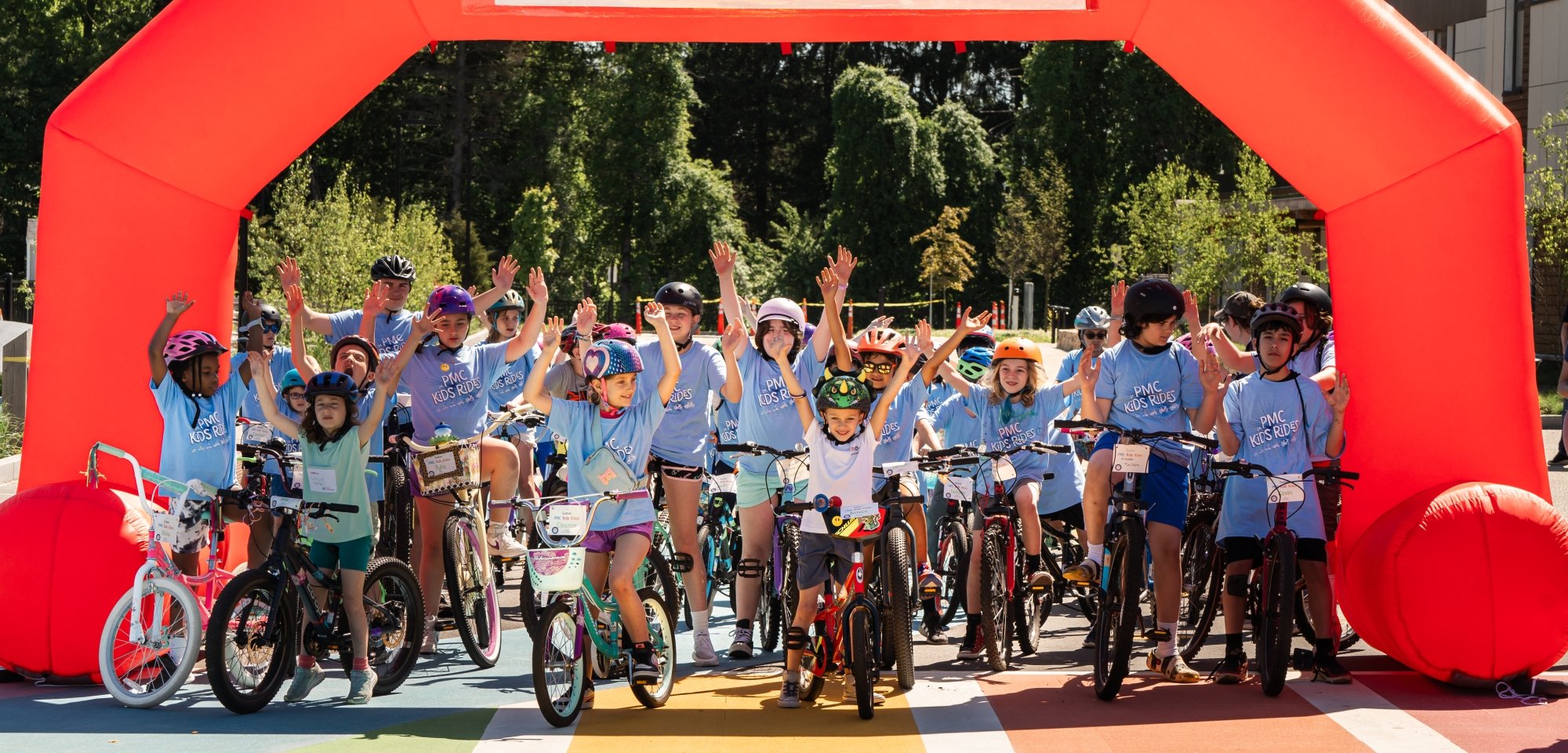 Group of young bike riders stand with arms in the air under 'PMC Kids Ride' start line