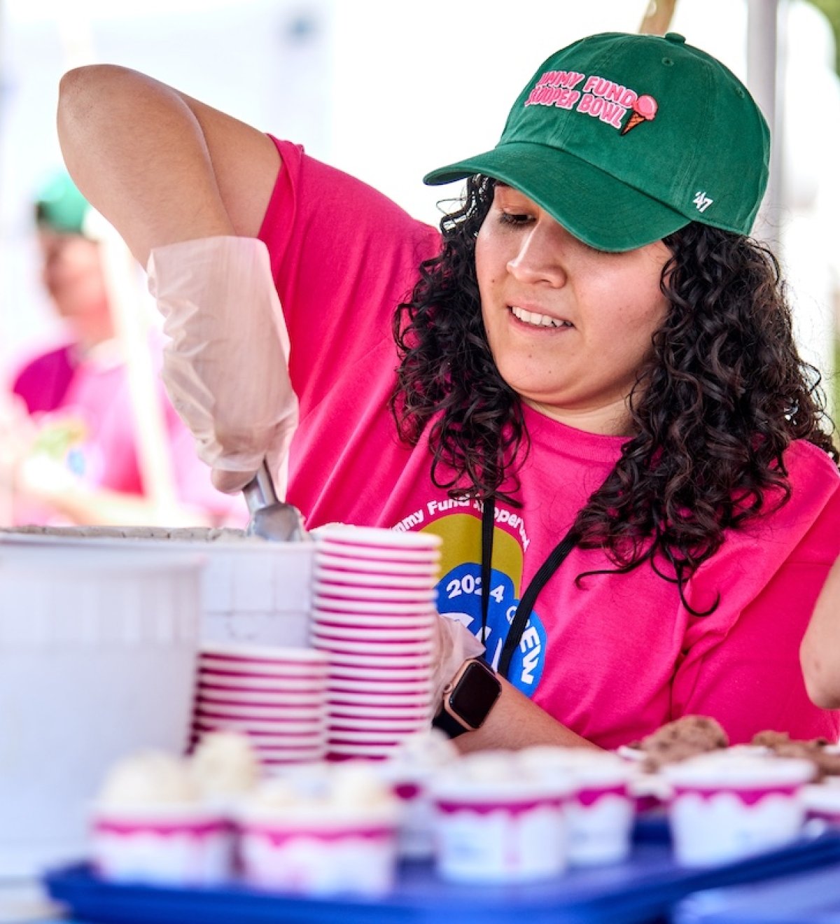 A volunteer in a bright pink Scooper Bowl T-shirt scoops ice cream out of a large tub