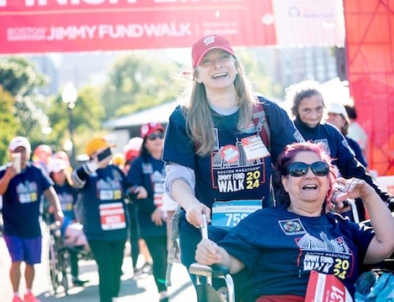 Two participants cross the finish line of the Jimmy Fund Walk; one participant is pushing the other in a wheelchair