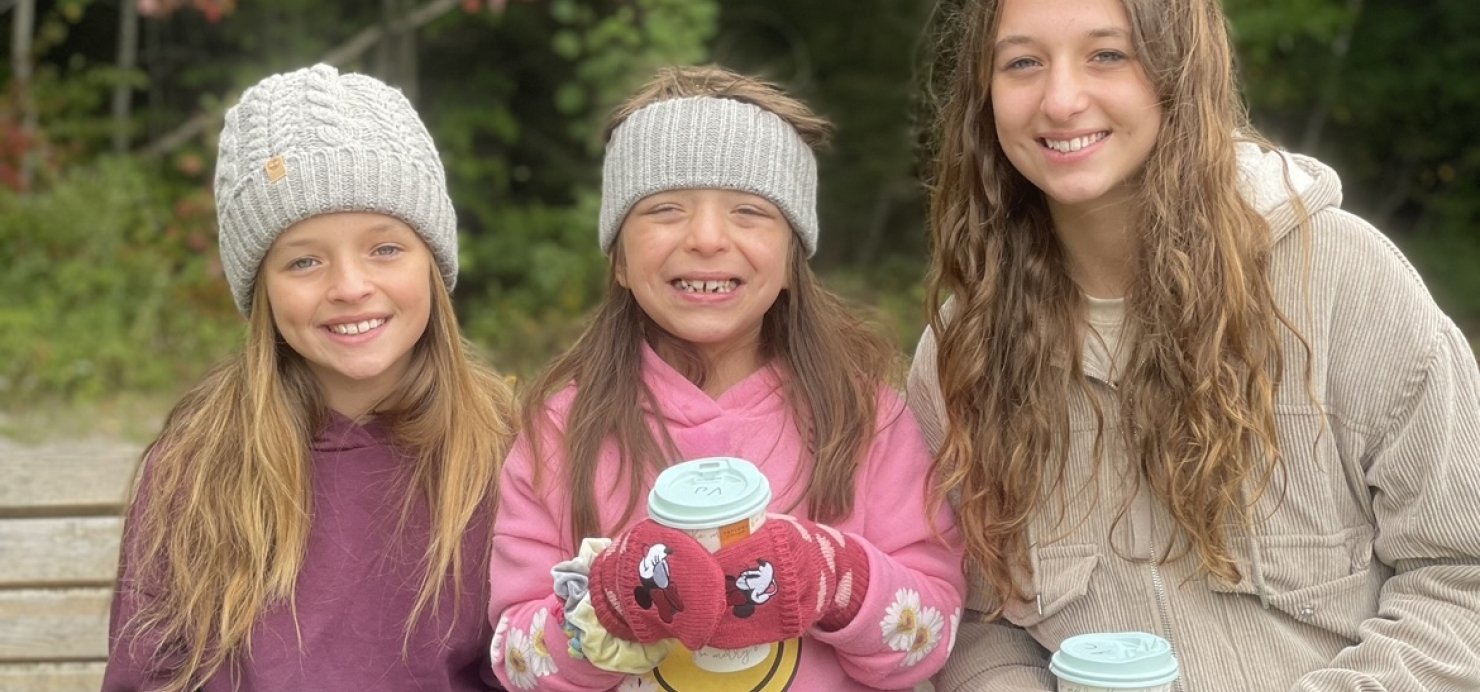 Three sisters sit on bench holding drinks with trees in background