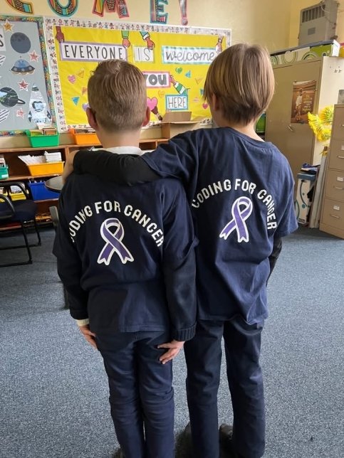 Two students in a classroom wearing Coding for Cancer shirts