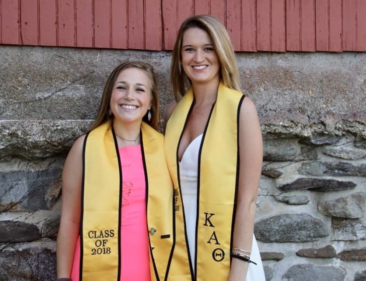 Grace and Gabby stand together in front of a wall wearing graduation clothes