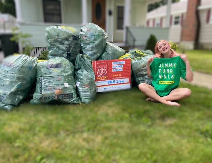 MacKenzie sits on lawn next to bags of cans holding up a 'Jimmy Fund Walk' T-shirt