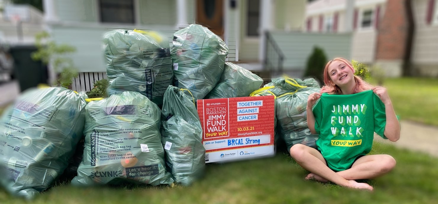MacKenzie sits on lawn next to bags of cans holding up a 'Jimmy Fund Walk' T-shirt