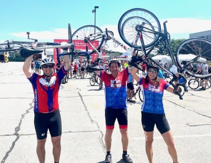 Three bikers stand together smiling and holding bikes above heads