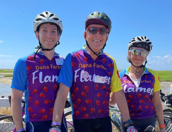 Three bike riders pose in front of a marsh while wearing helmets and 'Dana-Farber Flames' jerseys