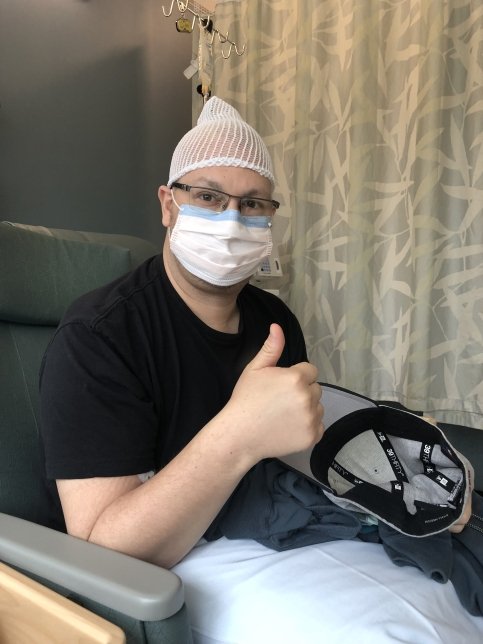 Matt sits in hospital chair wearing mask and giving a thumbs up