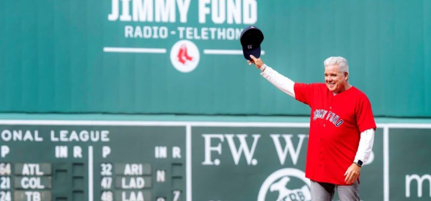 Doug stands on Fenway Park baseball field holding hat in air
