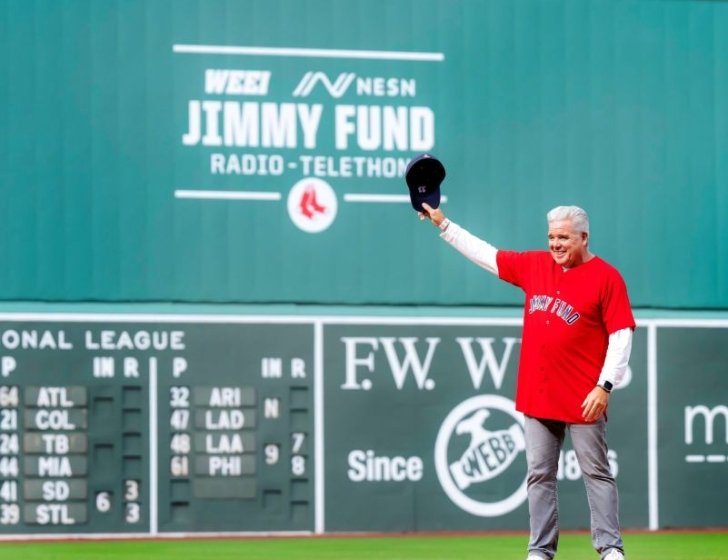 Doug stands on Fenway Park baseball field holding hat in air